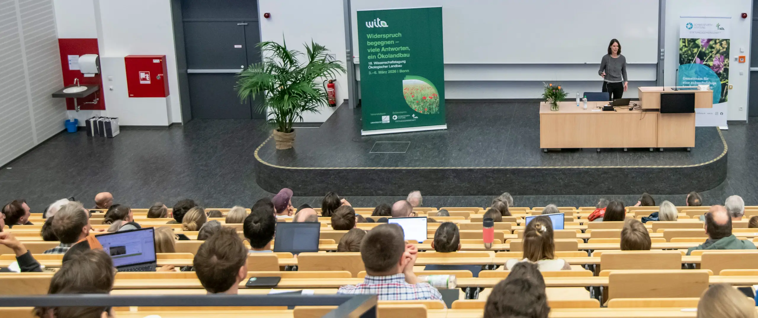 In einem Vorlesungssaal spricht eine Professorin mit schulterlangen dunklen Haaren und einem grauen Pullover zu ihren Zuhörern und Zuhörerinnen.